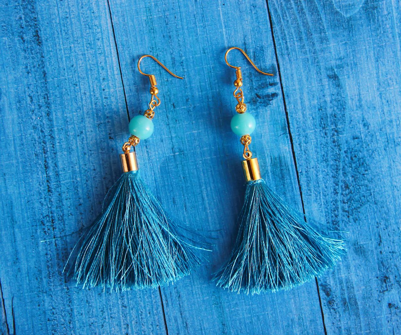 gallery-01 Stylish blue tassel earrings with gold accents displayed on a wooden surface.